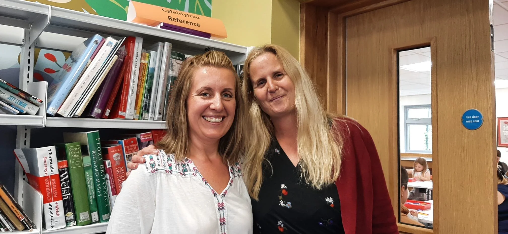 Two women standing in a library near bookshelves, smiling at the camera. One woman has shoulder-length blonde hair and is wearing a white blouse with embroidered details. The other woman has long blonde hair and is wearing a black top with floral patterns and a red cardigan. In the background, there are children sitting at tables through a window in a classroom.
