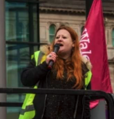 A woman speaking into a microphone during a protest, wearing a yellow vest, with a pink banner in the background.