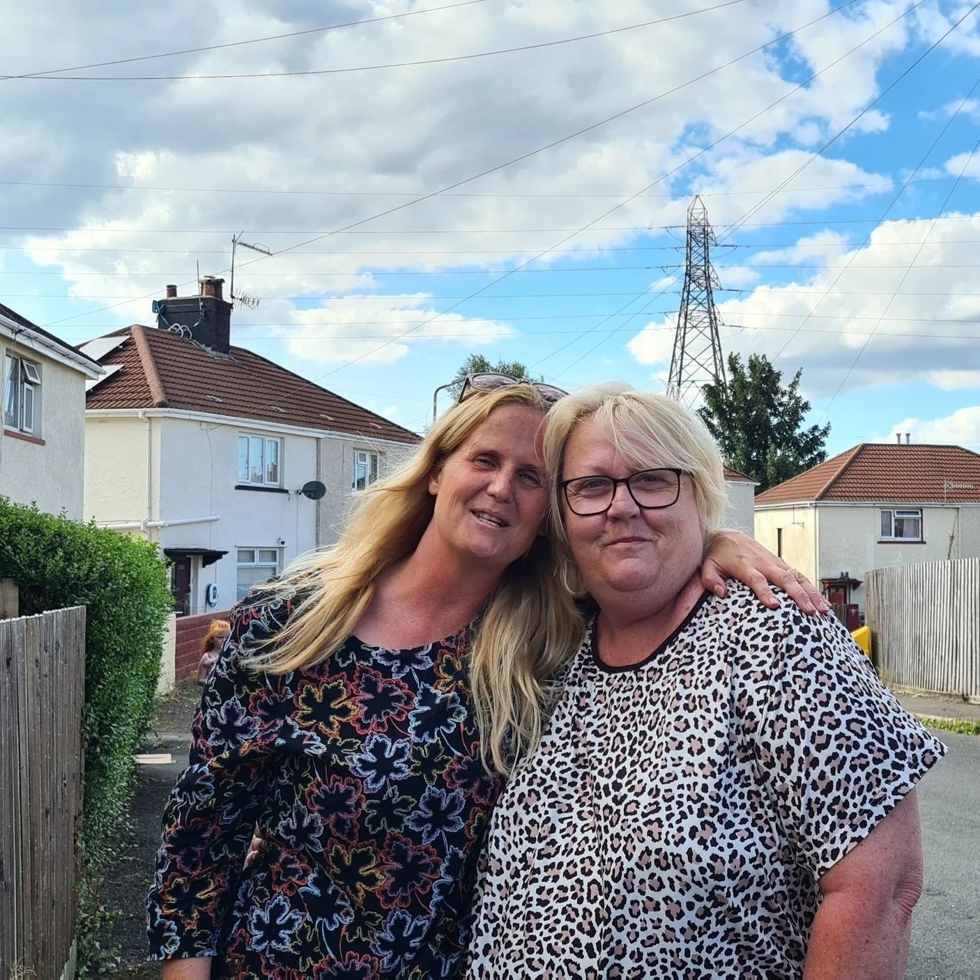 Two women standing outdoors, one with long blonde hair and the other with short blonde hair and glasses, posing in a neighborhood with houses, fences, and a large power line tower in the background.
