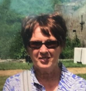A woman with short brown hair and sunglasses smiling outdoors near trees and a stone wall.