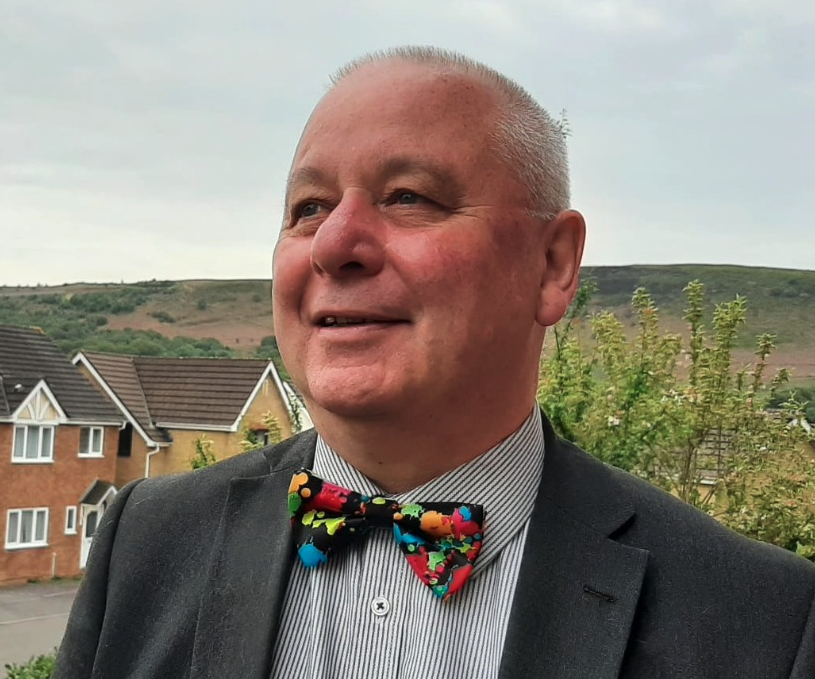 A smiling middle-aged man wearing a suit and a colorful, patterned bow tie, outdoors with residential houses and green hills in the background.