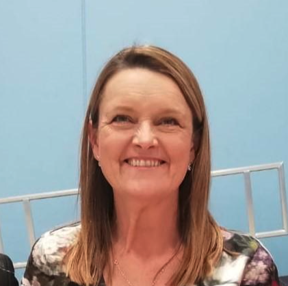 A woman with shoulder-length brown hair smiling, wearing a patterned shirt, sitting against a light blue wall.