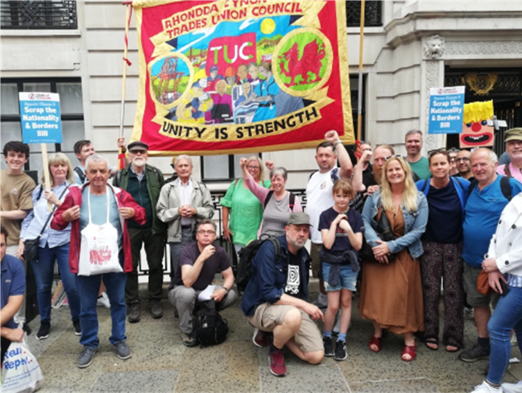 A group of people standing in front of a banner that reads 'Rhonda & Yvonne Trades Union Council, TUC, Unity is Strength.' The banner has colorful images and symbols representing unity, including two dragons and a globe. Some people are holding signs and flags, and the group appears to be participating in a protest or demonstration.