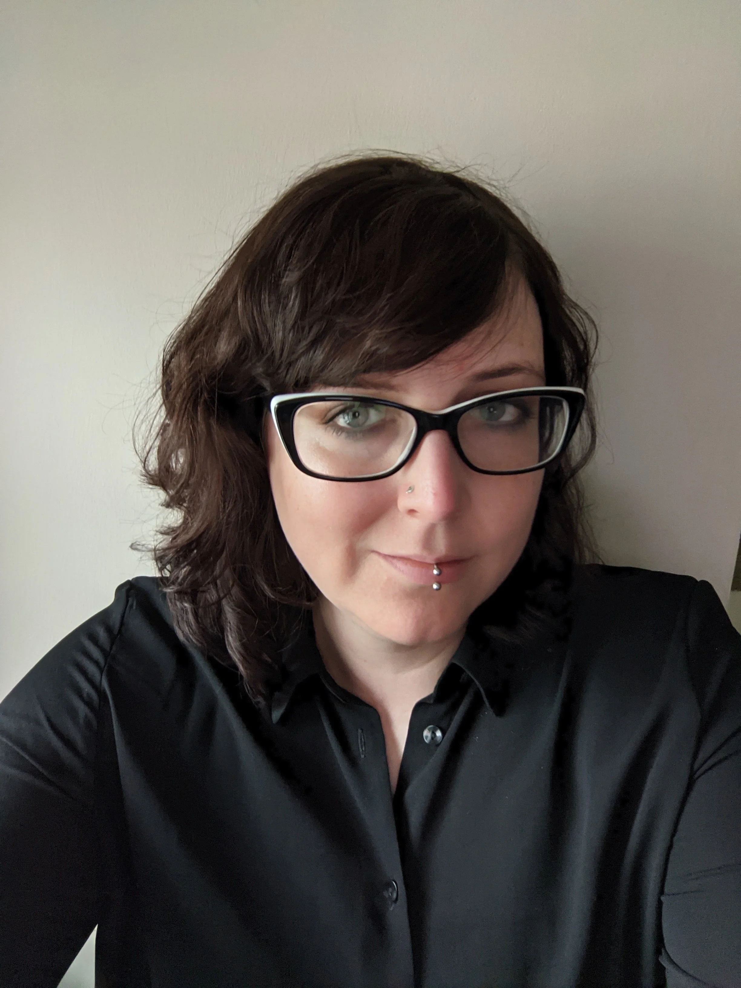 A woman with dark brown wavy hair, glasses, and piercing on her nose and lip, wearing a black button-up shirt, taking a selfie indoors against a plain white wall.