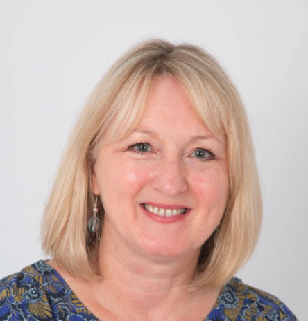 A woman with shoulder-length blonde hair, smiling, wearing a blue patterned top and earrings against a plain white background.