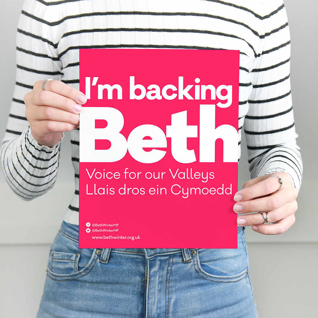 Person in a striped long sleeve shirt holding a pink campaign poster that reads, "I'm backing Beth," with additional text and social media details.