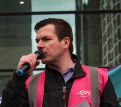 Man speaking into a microphone outdoors, wearing a pink backpack and a black jacket