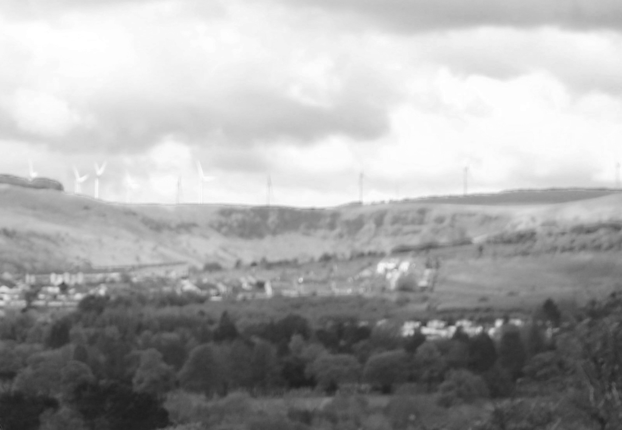 Black and white landscape of rolling hills with wind turbines on top, scattered houses in the middle ground, and trees in the foreground, under a cloudy sky.