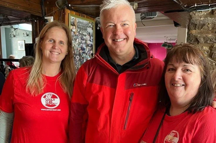 Three people smiling for a photo inside a restaurant or cafe, wearing red shirts and jackets, with a mantle and a framed collage of photos in the background.