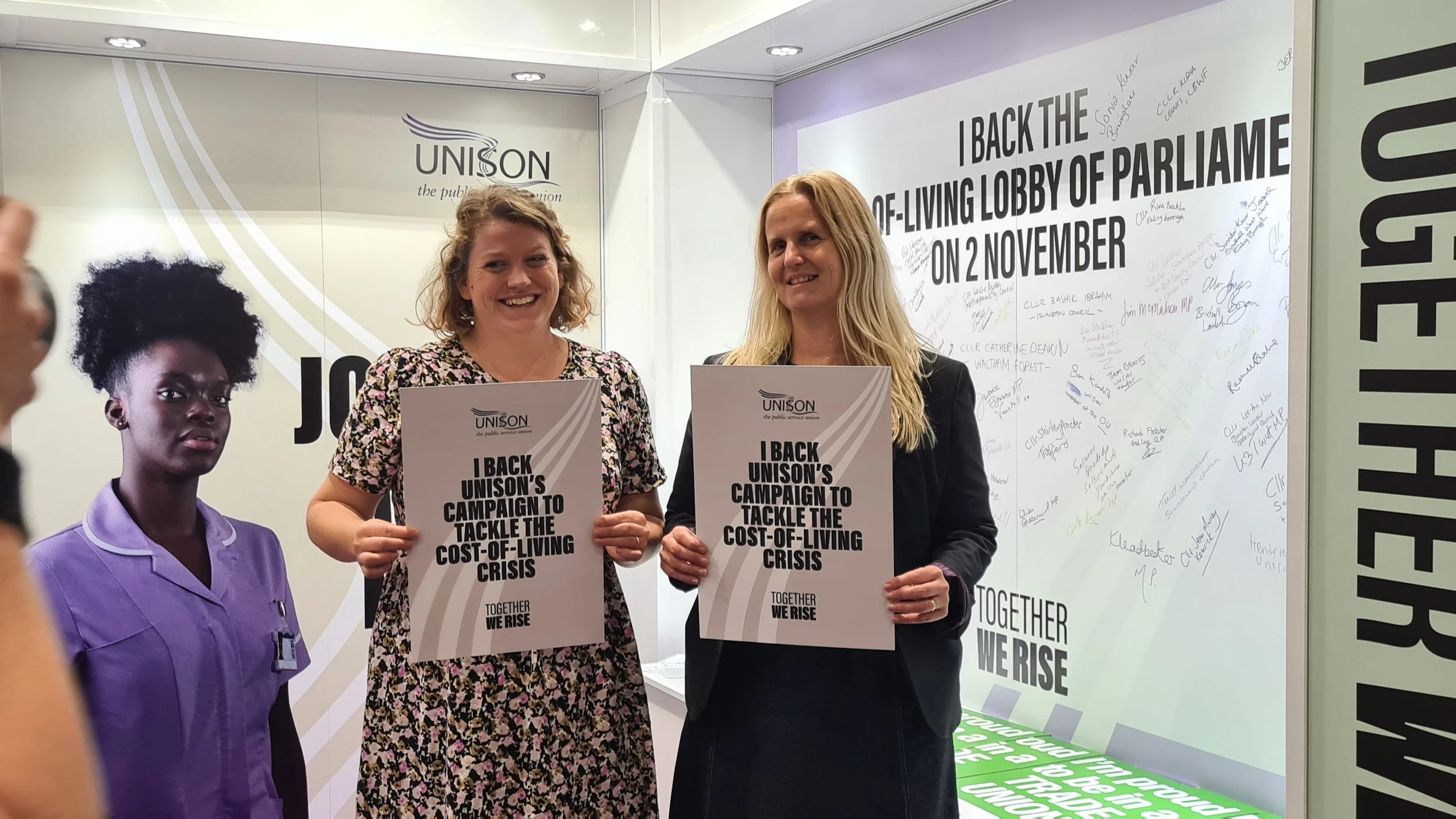 Two women holding UNISON campaign posters that read "I BACK UNISON'S CAMPAIGN TO TACKLE THE COST-OF-LIVING CRISIS." They stand in front of a white wall with a large sign and signatures. To the left, part of a person in purple scrubs is visible.