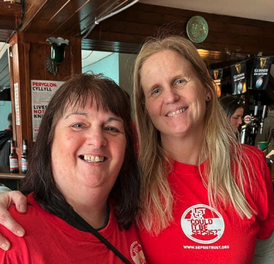 Two women smiling and wearing red shirts, standing close together inside a bar or pub.
