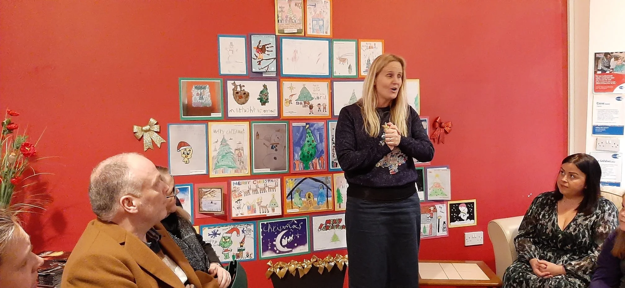 A woman with long blonde hair standing and speaking in front of a red wall decorated with Christmas artwork. Several people are seated listening, including an older man with glasses and a woman with dark hair.