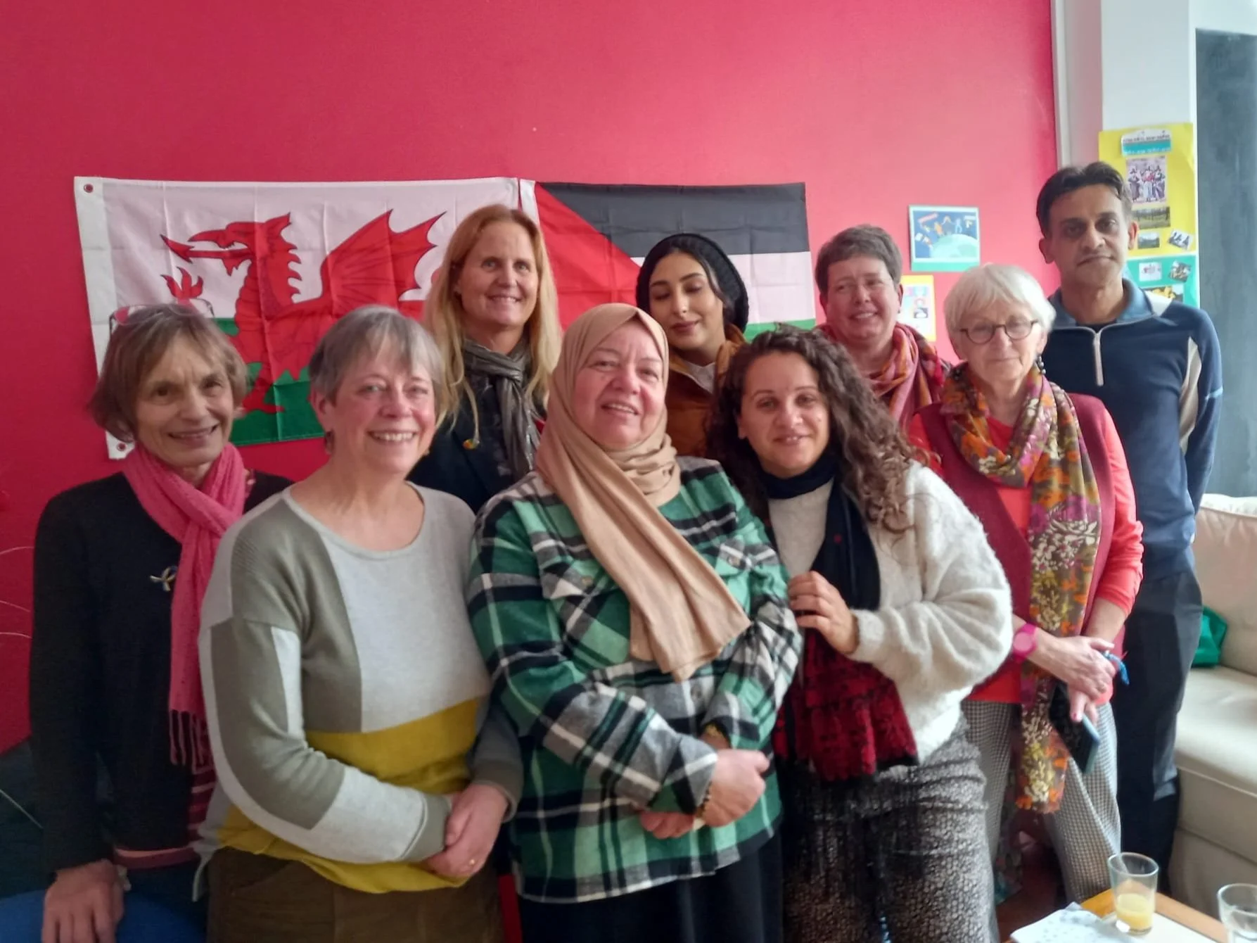 Group of ten people smiling, standing in front of a Welsh flag. The group includes both men and women of various ages. Some women are wearing scarves and sweaters. The background wall is painted red with some colorful posters and artwork, and there is a white sofa to the right.