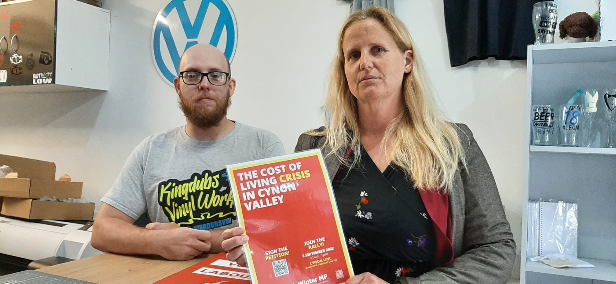 Two people sitting at a table in a room with a Volkswagen logo in the background. The person on the left is a man with glasses and a beard, wearing a gray T-shirt that says "Kingdubs Vinyl Worx." The person on the right is a woman with long blond hair, wearing a dark dress with floral patterns and a gray blazer, holding a red and yellow flyer about a rally in Clydon Valley.