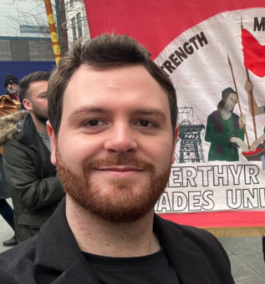 Smiling man with short dark hair and beard, standing outdoors with people and a red march banner in the background.