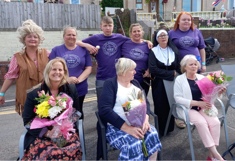 Group of women and girls gathering outdoors, some holding bouquets of flowers, in front of houses with a gray wall and a wooden fence.