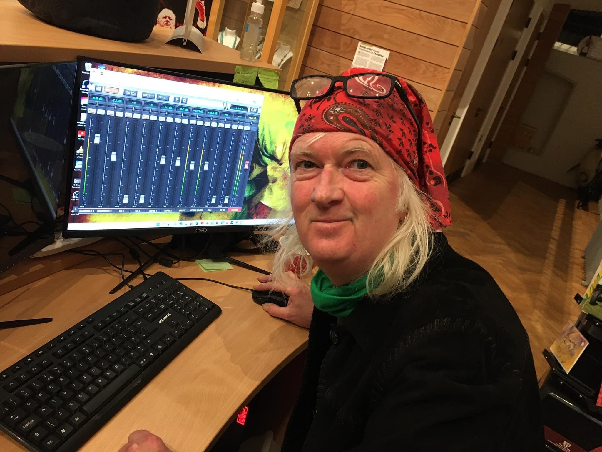 A man with long white hair, wearing a red bandana, glasses on his forehead, and a green shirt, operating a digital audio workstation with a large monitor, keyboard, and mouse in a wooden interior space.