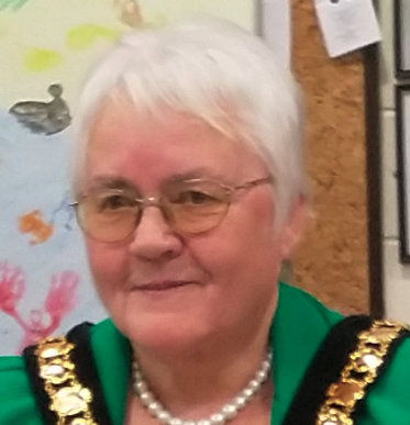 A woman with short white hair and glasses, wearing a green top with gold accents and a pearl necklace, standing in front of a colorful child's drawing and wooden corkboard.