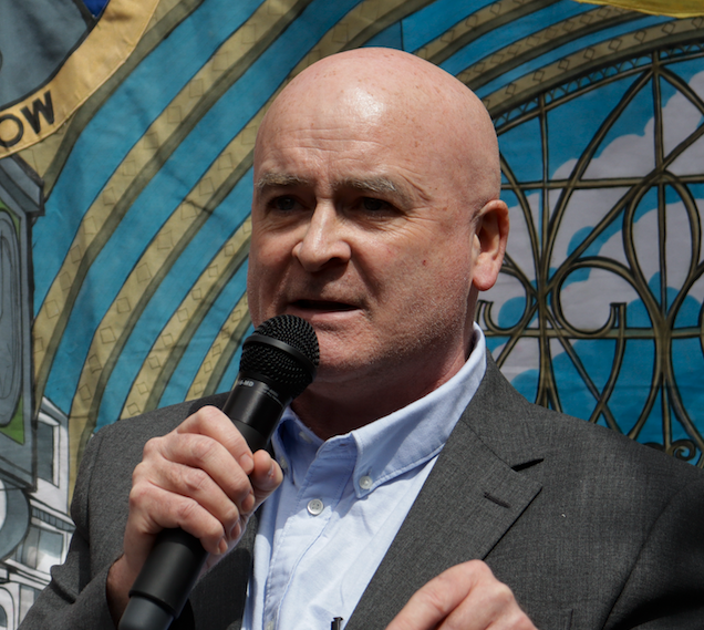 A bald man in a suit speaking into a microphone, standing in front of a colorful mural background.