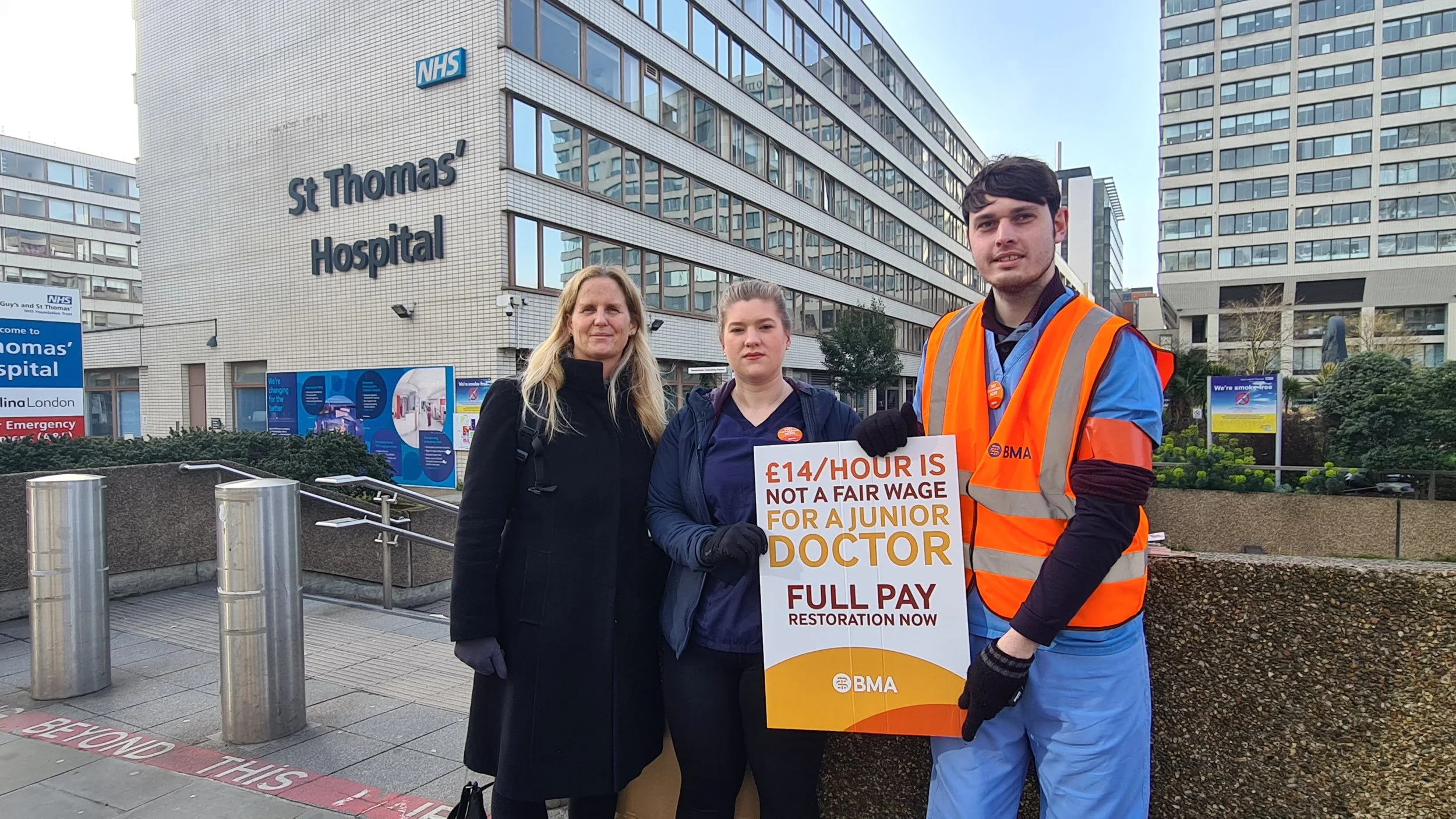 Three people, including a woman in black coat and a person in medical scrubs, standing outside St. Thomas' Hospital in London, holding a protest sign about fair wages for junior doctors.