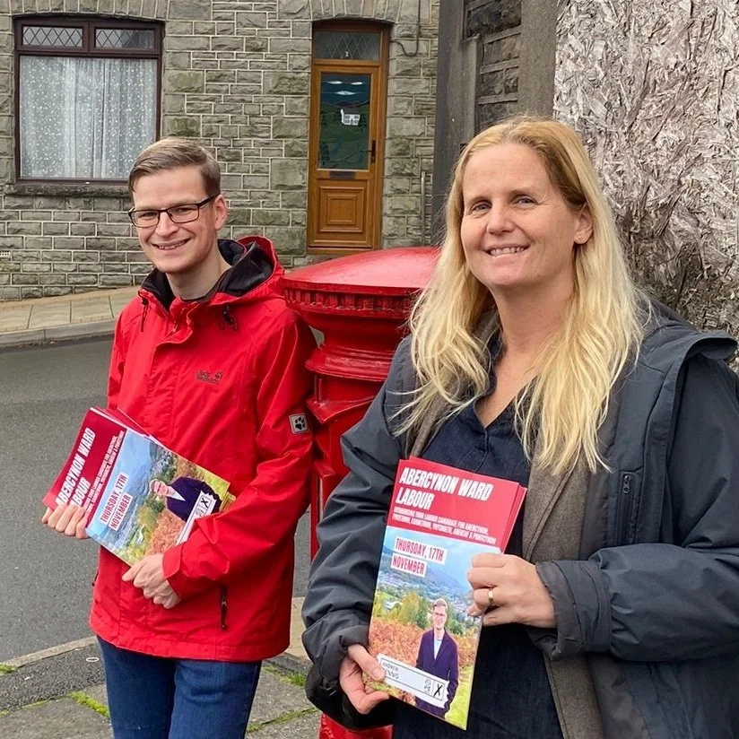 Two people standing on a sidewalk holding flyers, with an old stone building and a red post box behind them. The man is wearing glasses and a red jacket, and the woman has long blonde hair and a black jacket.