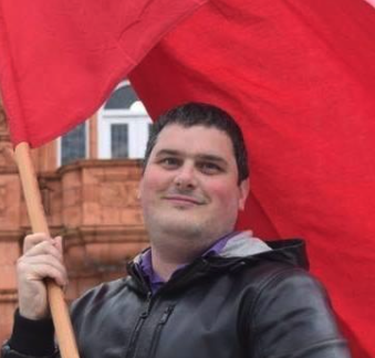 A person holding a red flag on a wooden pole, smiling outdoors with a building in the background.