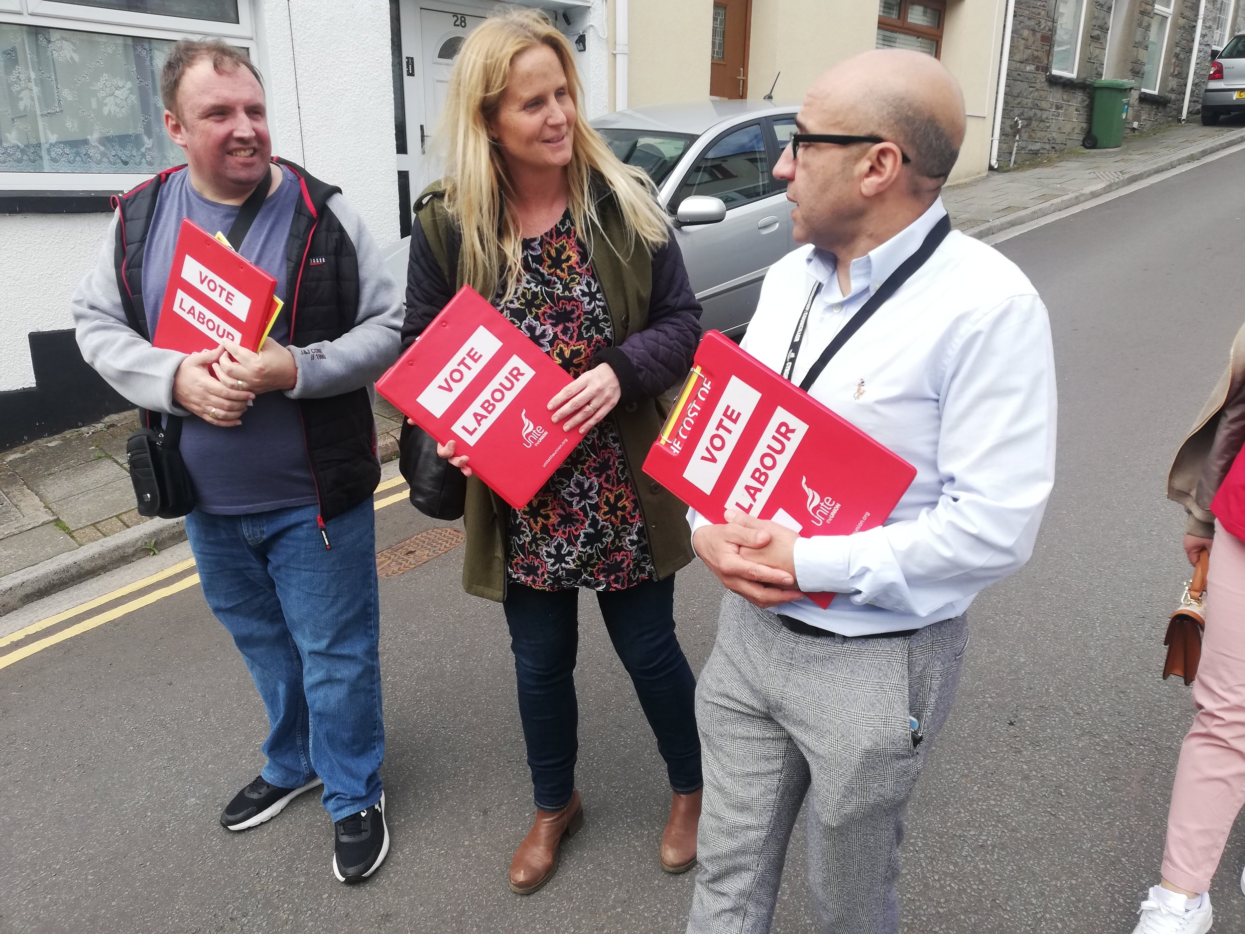 Three people standing on a sidewalk, holding red campaign signs that read "Vote Labour" with the Labour party logo, engaging in conversation during a political campaign.
