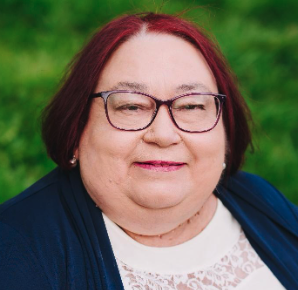 Close-up of a woman with red hair, glasses, and a slight smile, outdoors with green foliage in the background.