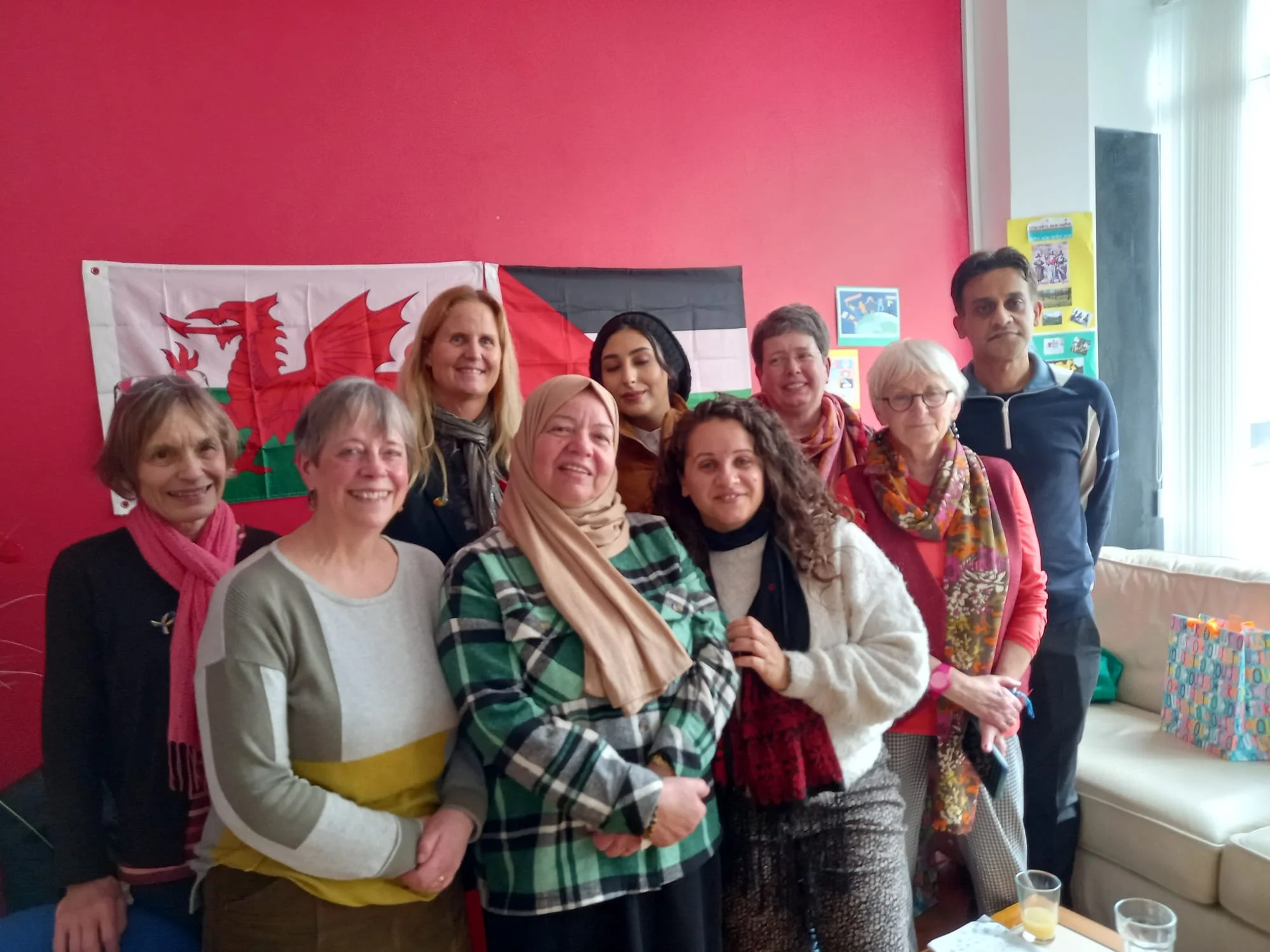 A diverse group of nine women and one man standing together indoors in front of a red wall with Welsh and German flags. They are smiling and appear to be celebrating or gathering for a social event.