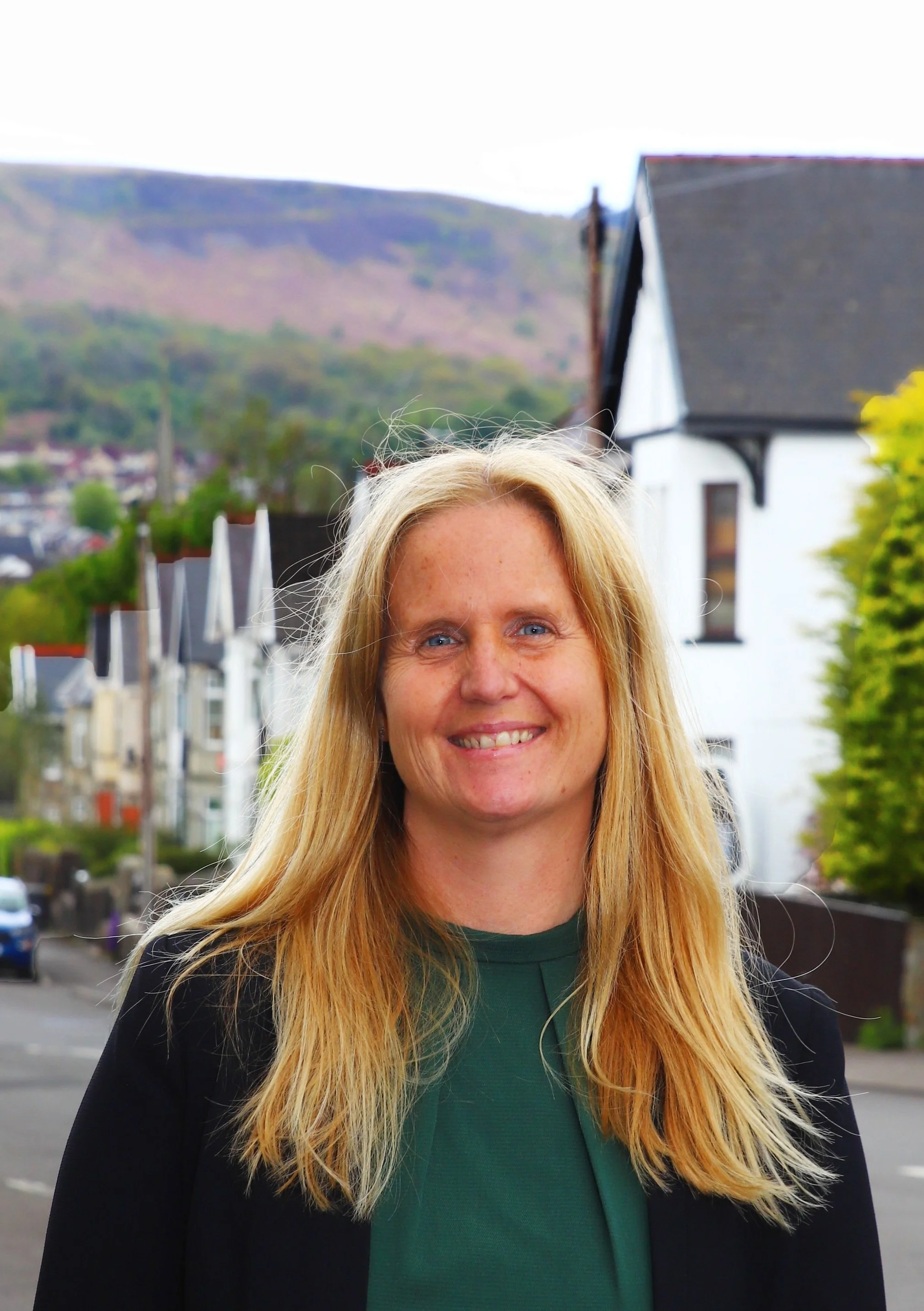 A woman with long blonde hair and a green top standing outdoors in a residential neighborhood with houses, a street, and hills in the background.