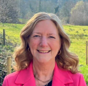 A smiling woman with blonde wavy hair wearing a pink blazer outdoors in a green landscape with trees and grass.