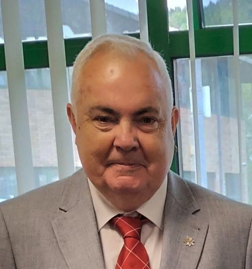 A man dressed in a gray suit, white shirt, and red tie, standing indoors in front of a window with green framing and vertical blinds.
