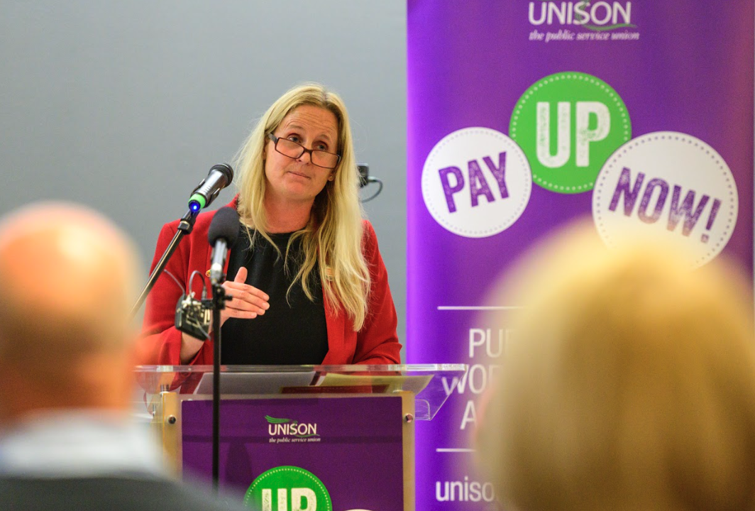 A woman with long blonde hair, glasses, and a red blazer speaking at a podium during a UNISON event, with a purple banner behind her that says "PAY UP NOW!" and mentions public service workers.