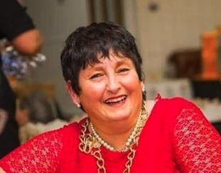 A woman with short dark hair wearing a red top and gold jewelry, smiling brightly at a social gathering.