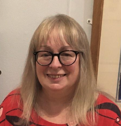 A woman with long blonde hair, glasses, and a red-and-black striped shirt smiling indoors.