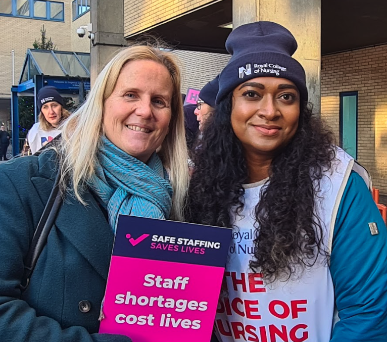 Two women outdoors, one holding a sign that reads 'Staff shortages cost lives,' during a rally or protest supporting nursing staff.