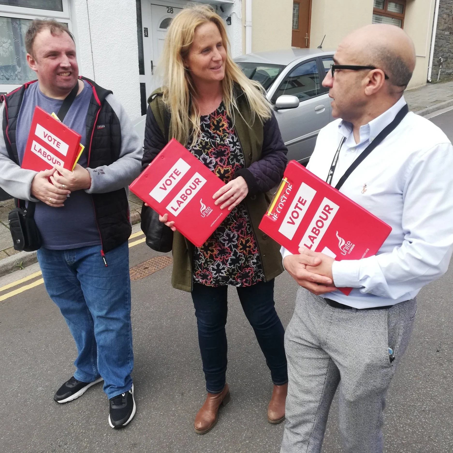 Three people standing outside talking, each holding a red folder that says 'VOTE LABOUR'. A man on the left has short hair and a gray hoodie, a woman in the middle has long blonde hair and a floral dress with a green jacket, and a bald man on the right is wearing glasses, a white shirt, and patterned pants.