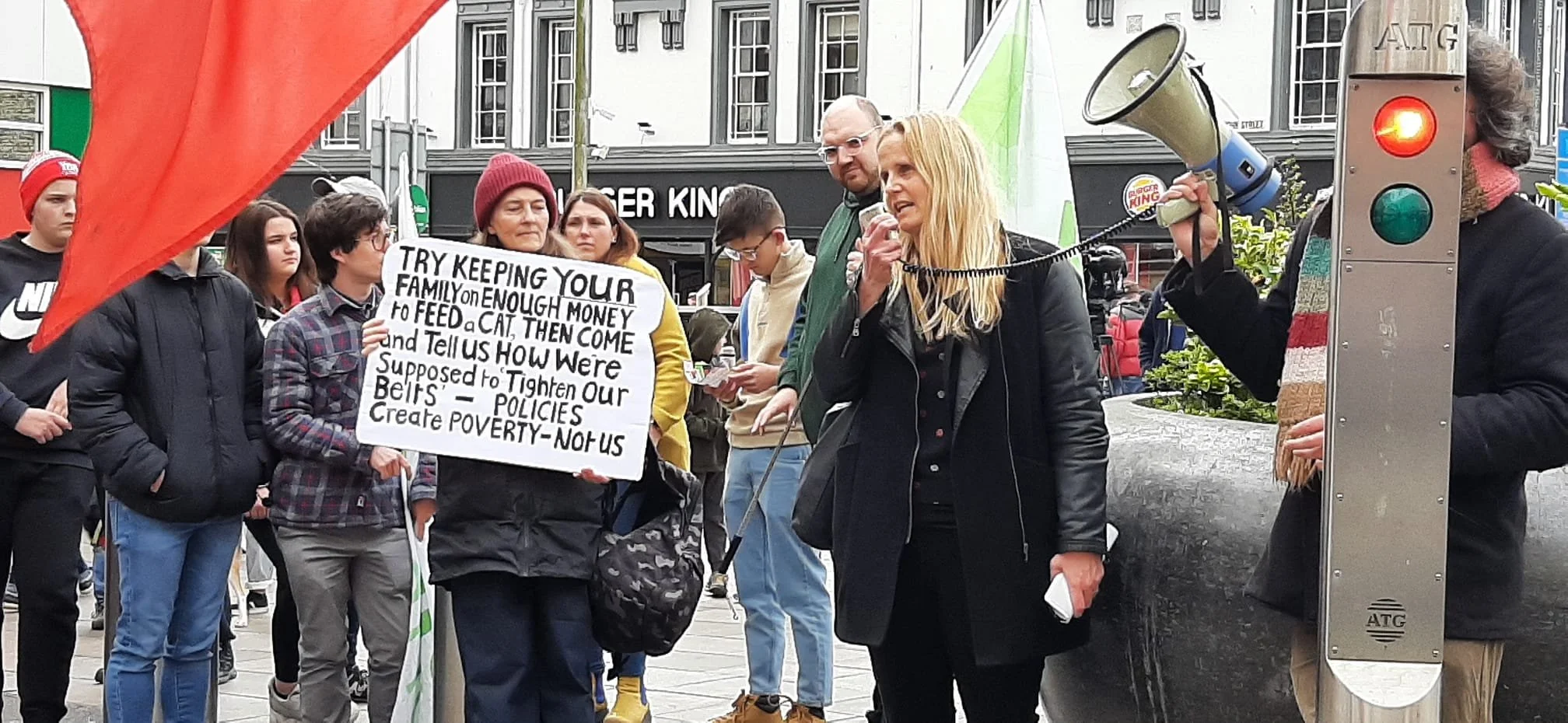 A woman speaking into a megaphone at a protest or rally, surrounded by people holding signs and banners on a city street.