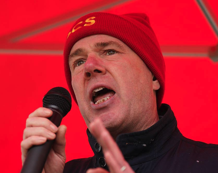 A man wearing a red beanie hat and black jacket speaking into a microphone, with a red background.