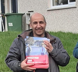 A smiling man holding a brochure or magazine outdoors in front of a house with green grass and a green trash bin behind him.