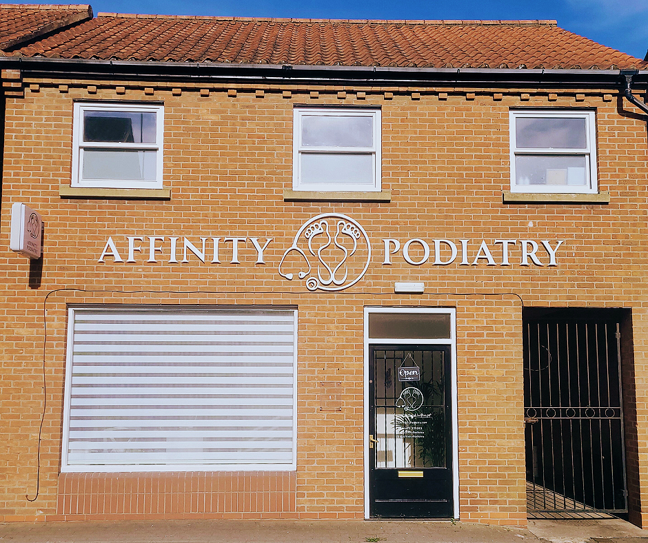 A brick building with three upper windows and a sign reading 'Affinity Podiatry' along with a logo of two interconnected feet. The entrance door has an 'Open' sign, and there is a large window with white blinds on the lower level.