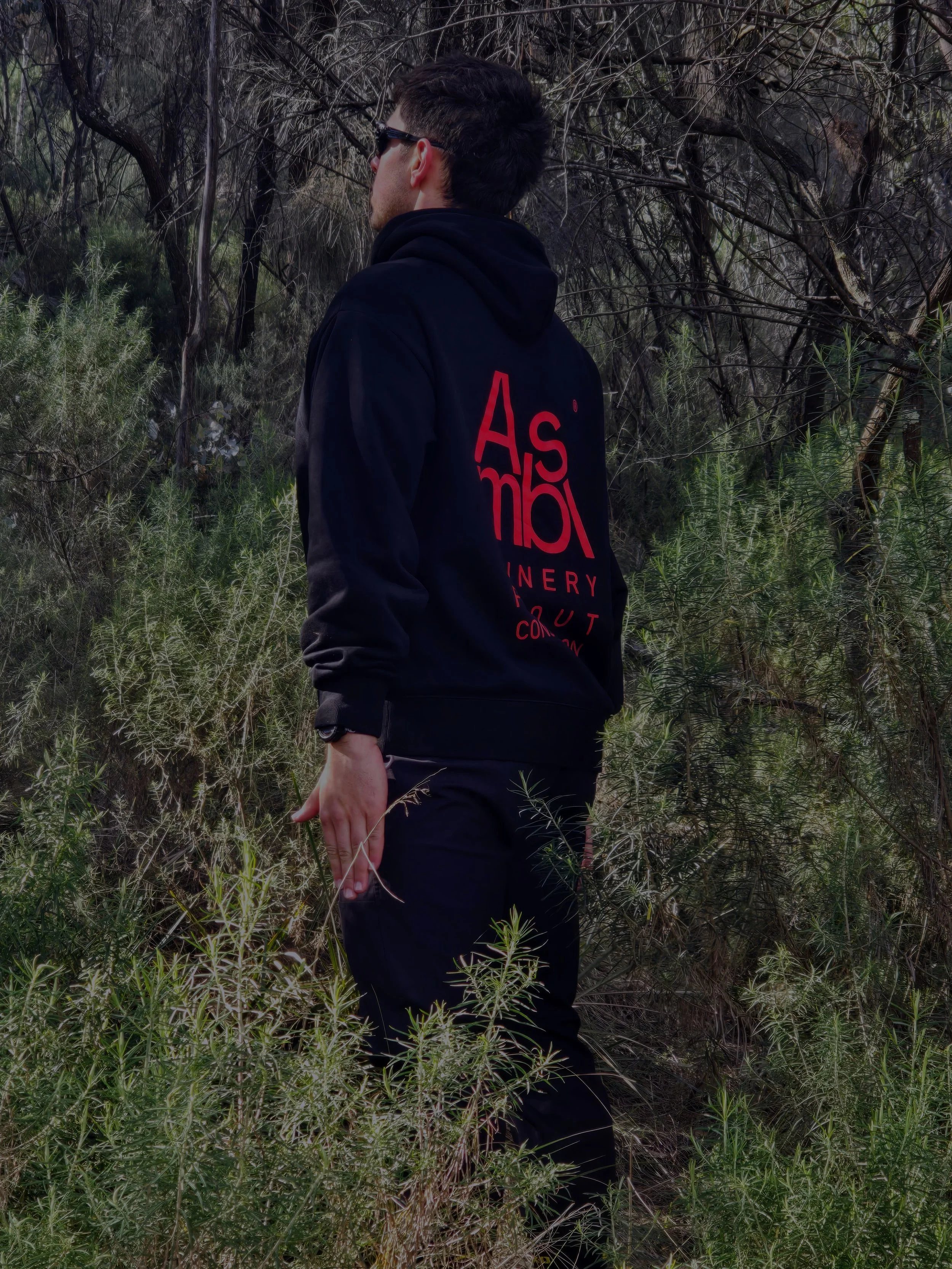 Man wearing black hoodie with red text standing in dense woods with green plants and trees around him, facing sideways.