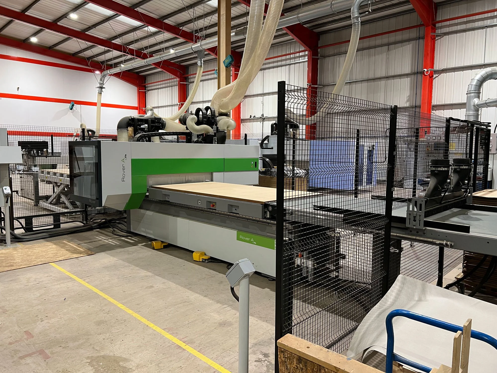 Industrial woodworking machine in a workshop with red, white, and gray walls, multiple hoses and ventilation systems, and safety fencing surrounding the equipment.
