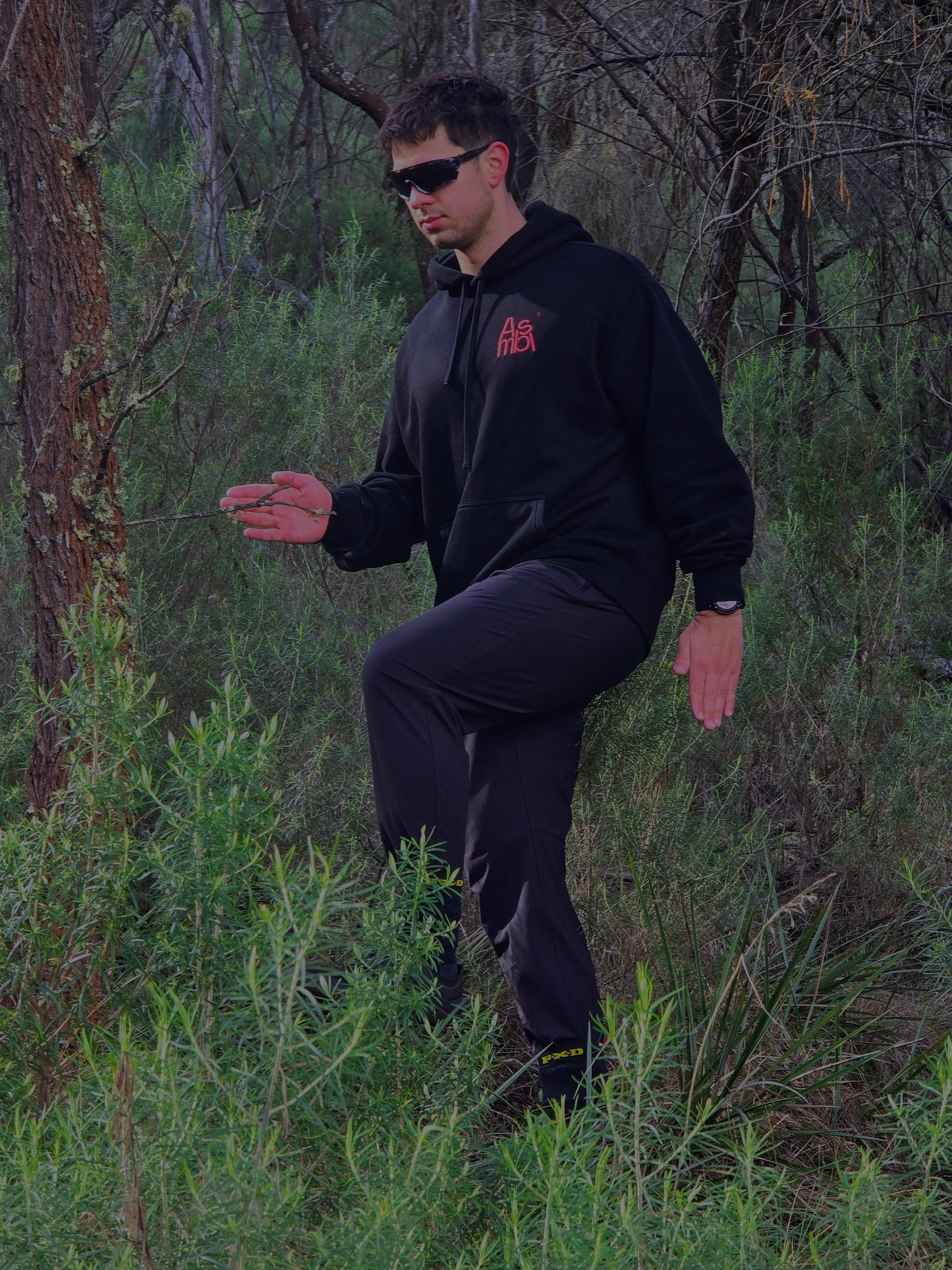 A young man with dark hair wearing sunglasses, a black hoodie, and black pants balances on one leg in a wooded area, holding a small branch in his right hand.