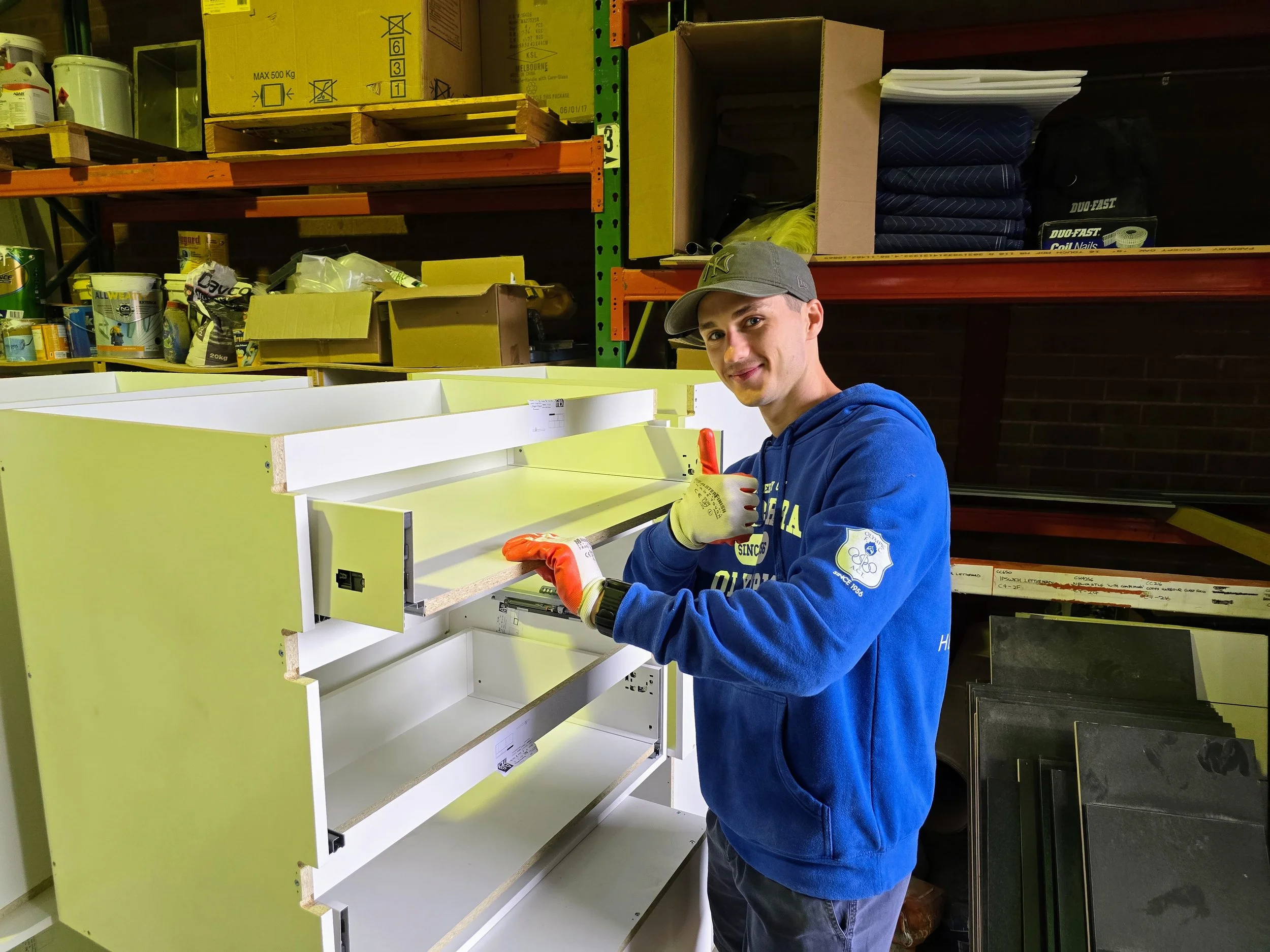 Young man wearing a gray cap, blue hoodie, and gloves assembling a white piece of furniture in a warehouse or hardware store. Shelves with various boxes and supplies are visible in the background.