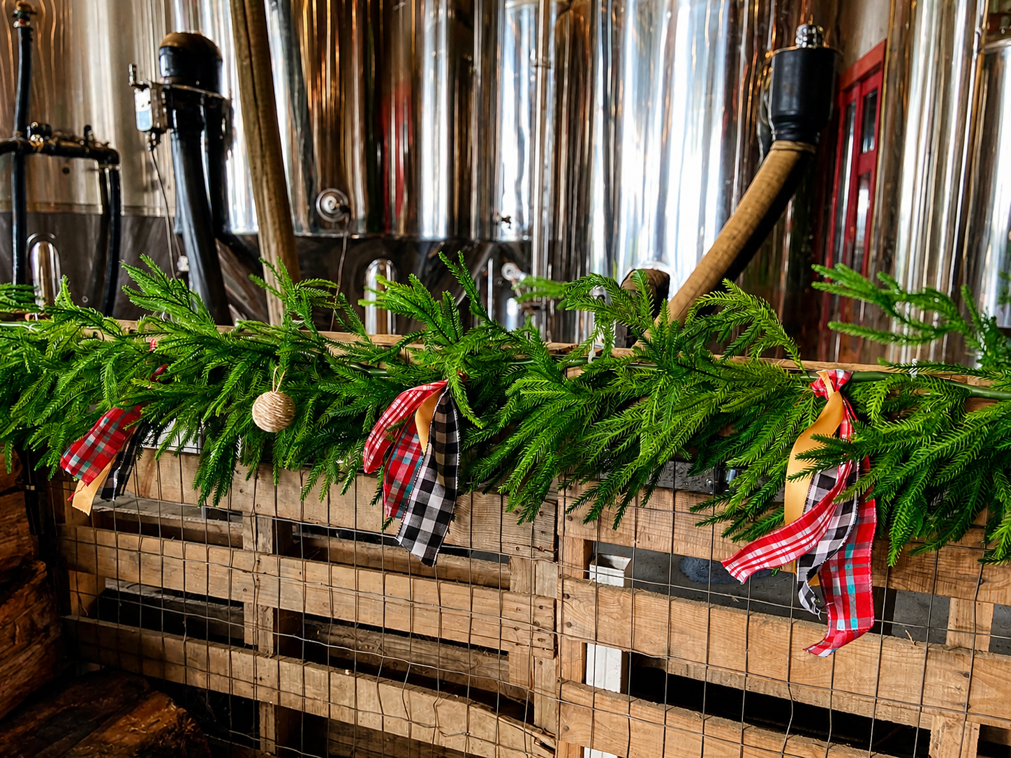 Fresh evergreen garland layered with plaid ribbon, neutral accents, and rustic textures adds seasonal warmth to the industrial brewery setting.