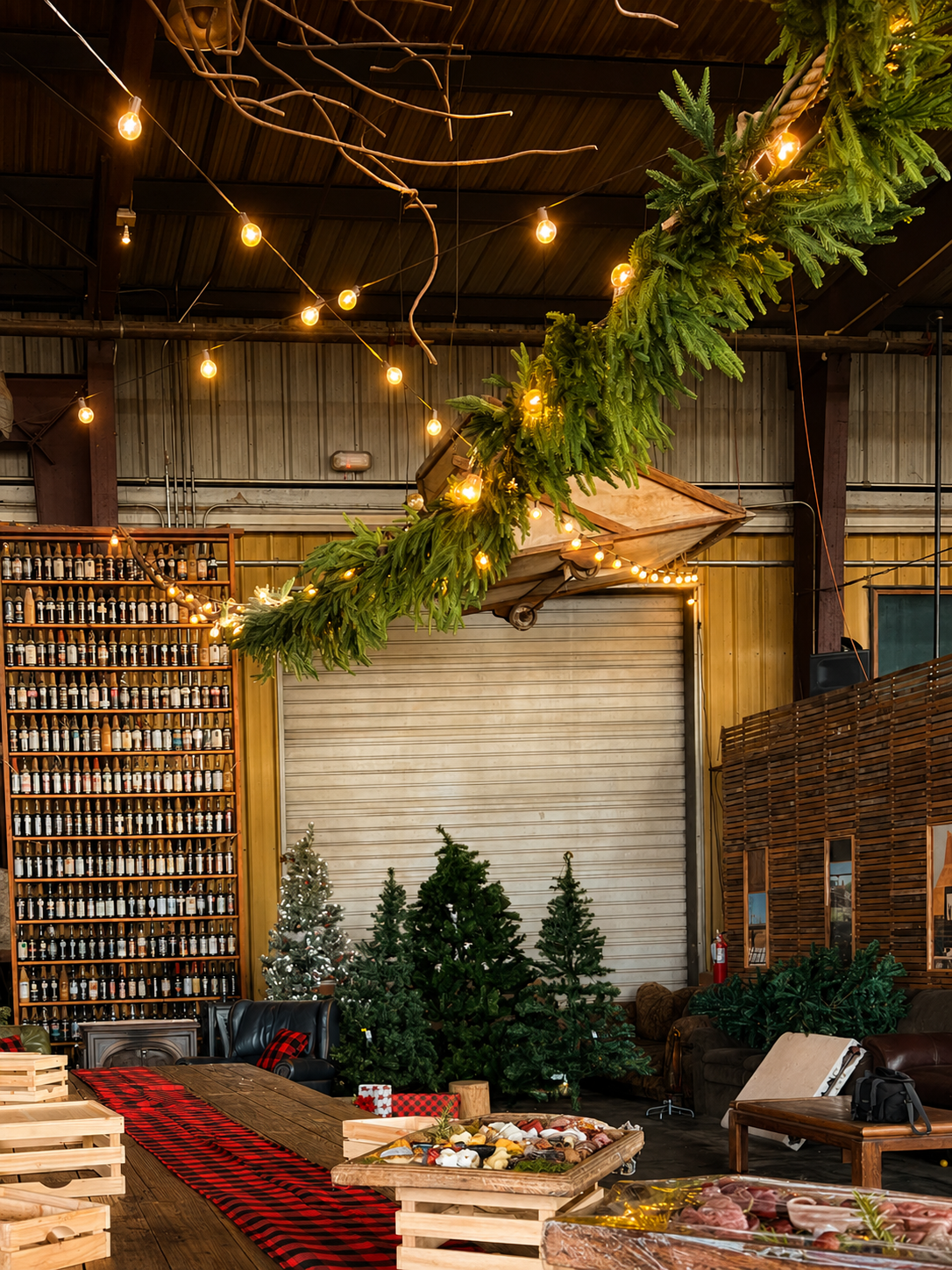 Overhead greenery, warm string lighting, Christmas trees, and rustic furnishings transform the industrial venue into a cozy and festive holiday celebration space.