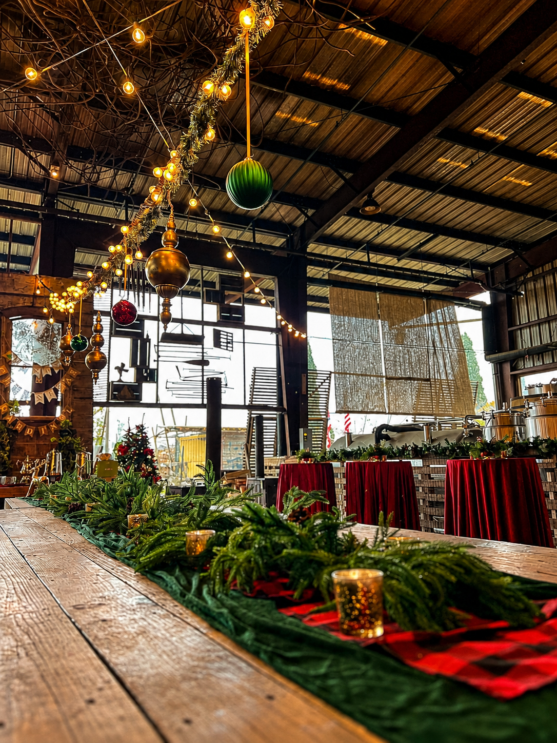Industrial holiday event venue styled with hanging ornaments, string lights, greenery and red cocktail tables.