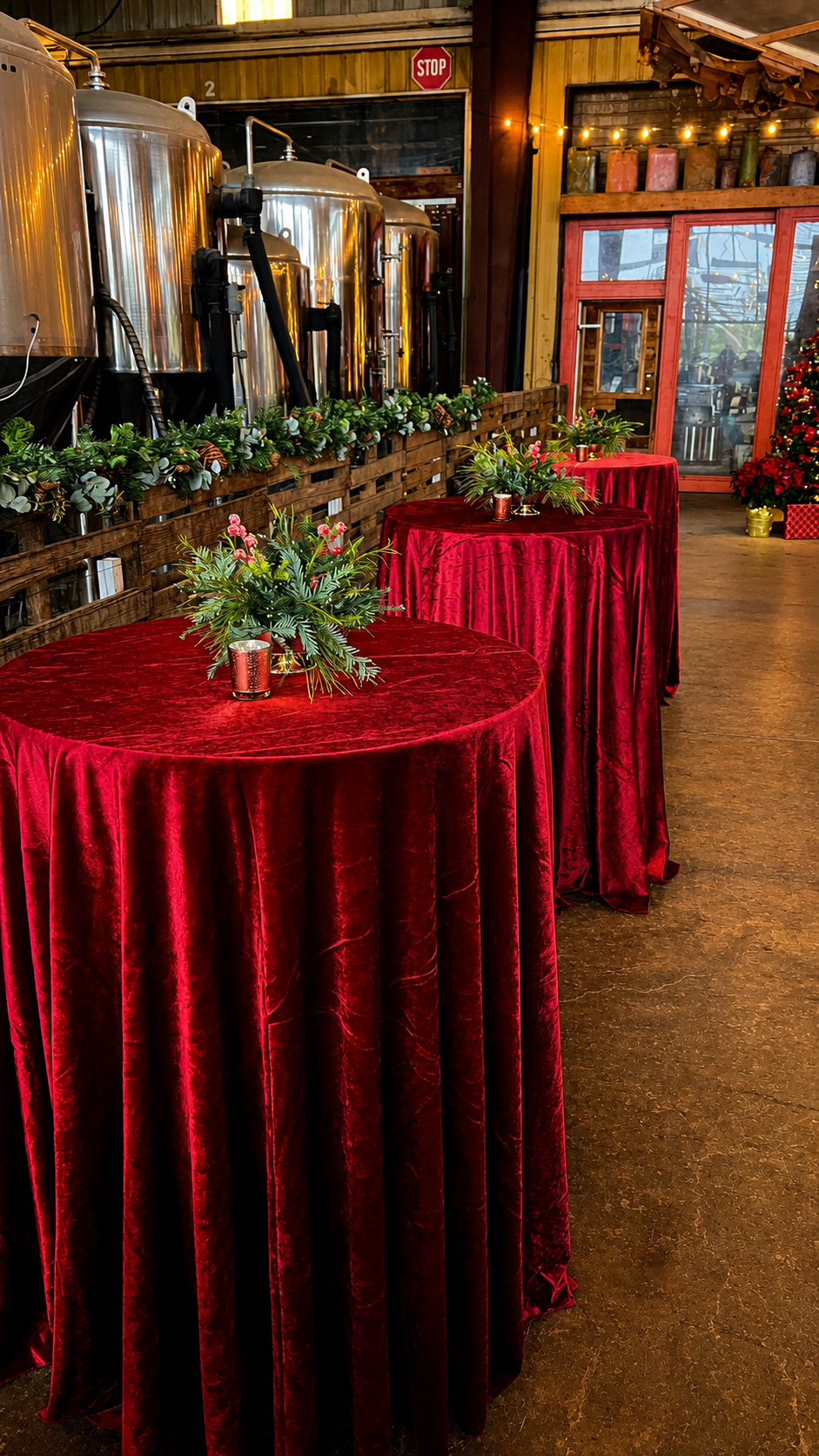 Red velvet cocktail tables with holiday greenery centerpieces inside a decorated industrial event venue.