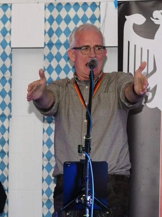 A man with short gray hair, glasses, wearing a checkered shirt and a rainbow-colored tie, holding a microphone in one hand and a green bottle in the other, speaking or singing at a microphone stand.